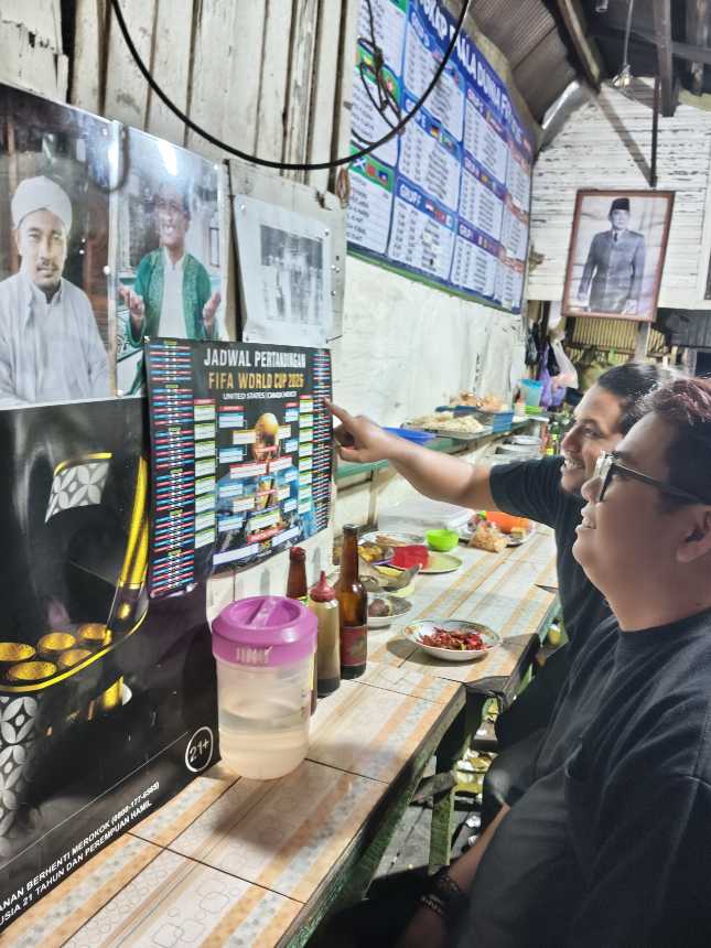 PANTAUJADWAL PIALA DUNIA – Arief dan Memet memperhatikan jadwal pertandingan Piala Dunia 2026 pada poster kecil sambil menikmati mi di Warung Kai Adi, Banjarmasin. Demam Piala Dunia mulai terasa di kalangan pencinta sepak bola yang bersiap begadang mendukung tim favorit mereka. (foto: Mercy )